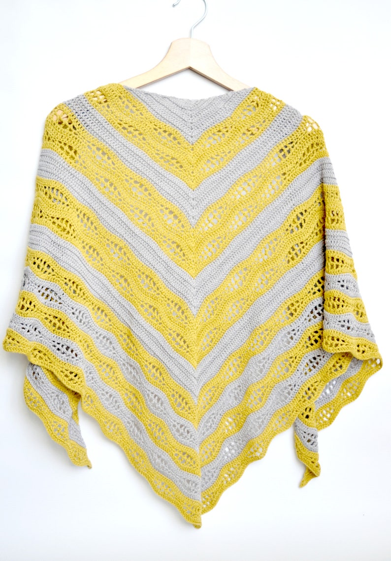 May include: A handmade, chevron-patterned shawl in yellow and gray. The shawl is made of yarn and is hanging on a wooden hanger against a white background. The design features alternating stripes of yellow and gray, with a lacy, openwork pattern.