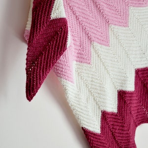 May include: A crocheted chevron shawl in shades of pink, white and burgundy. The shawl is made with a simple stitch pattern and has a triangular shape.