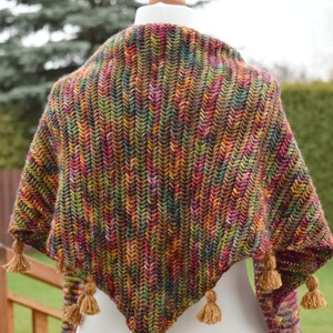 Puede incluir: Un chal triangular de ganchillo hecho con un hilo multicolor y jaspeado en tonos marrones, verdes, rojos y amarillos. El chal tiene una franja de borlas a lo largo del borde inferior.