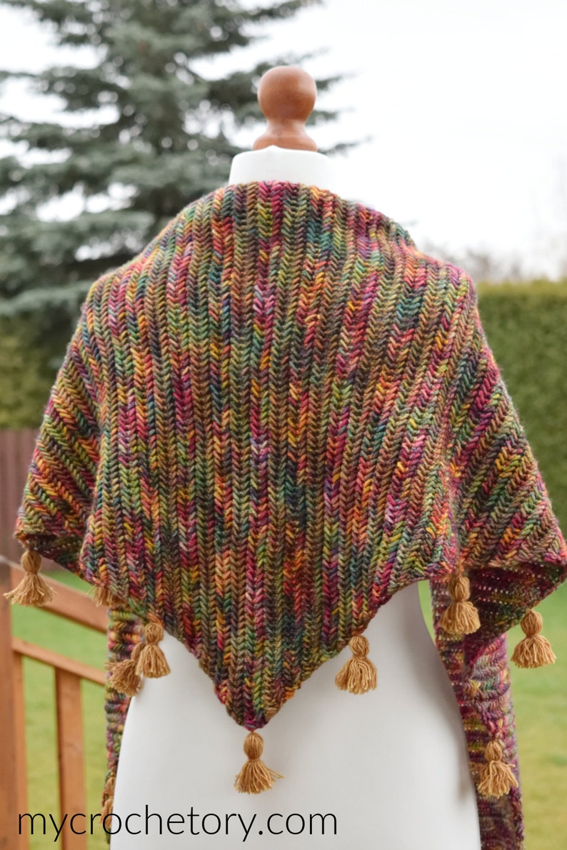 Puede incluir: Un chal triangular de ganchillo hecho con un hilo multicolor y jaspeado en tonos marrones, verdes, rojos y amarillos. El chal tiene una franja de borlas a lo largo del borde inferior.