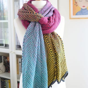 Puede incluir: Una bufanda de ganchillo colorida con un degradado de azul, amarillo y rosa. La bufanda está drapeada sobre un maniquí blanco y anudada en el cuello. La bufanda está hecha con un punto texturizado y tiene flecos en los extremos.