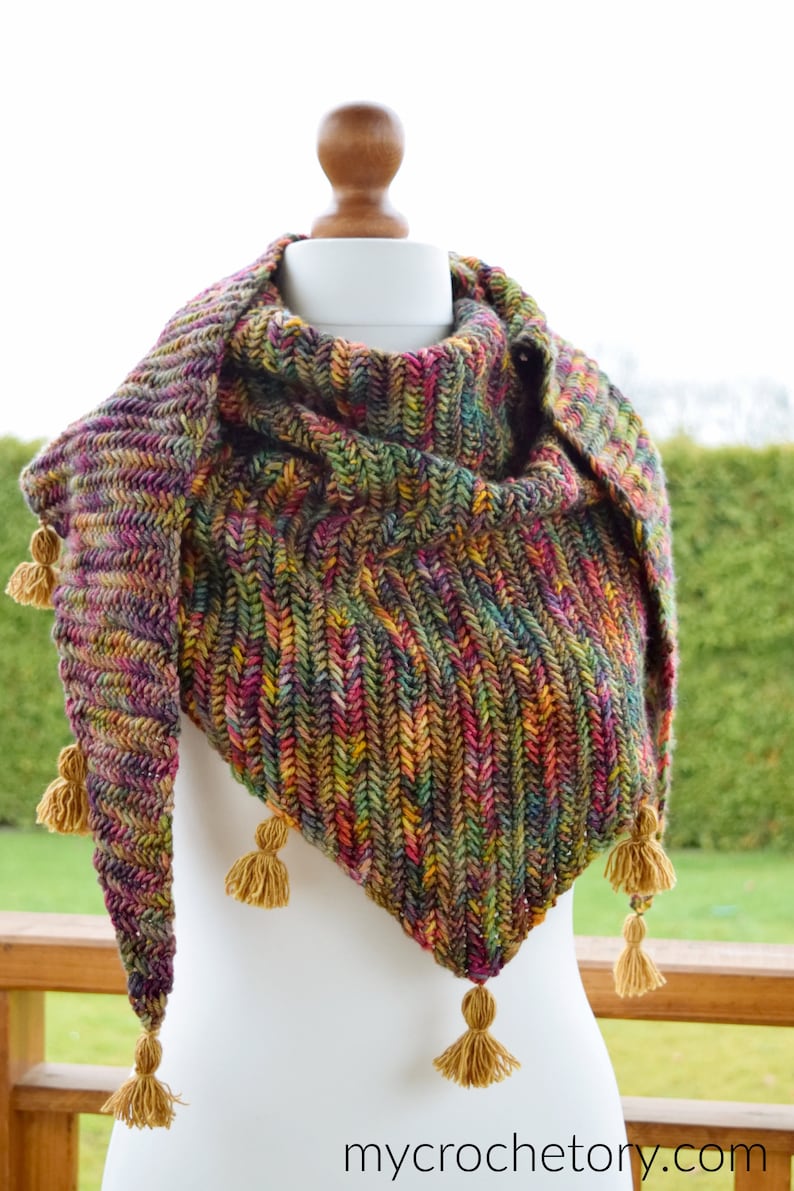 Puede incluir: Un chal triangular de lana multicolor con tonos de verde, marr&oacute;n, morado y rojo. El chal tiene un patr&oacute;n de punto texturizado y est&aacute; terminado con borlas doradas.