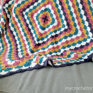 Könnte beinhalten: Eine bunte Häkeldecke mit einem quadratischen Muster in der Mitte. Die Decke ist aus verschiedenen Farben gefertigt, darunter Teal, Senfgelb, Pink, Weiß und Grau. Die Decke ist gefaltet und über ein graues Sofa gelegt.