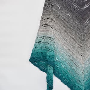 May include: A handmade, open-weave shawl in a gradient of colors, from dark gray to teal. The shawl is made of soft yarn and is draped over a wooden hanger. The intricate crochet pattern is visible throughout the design.