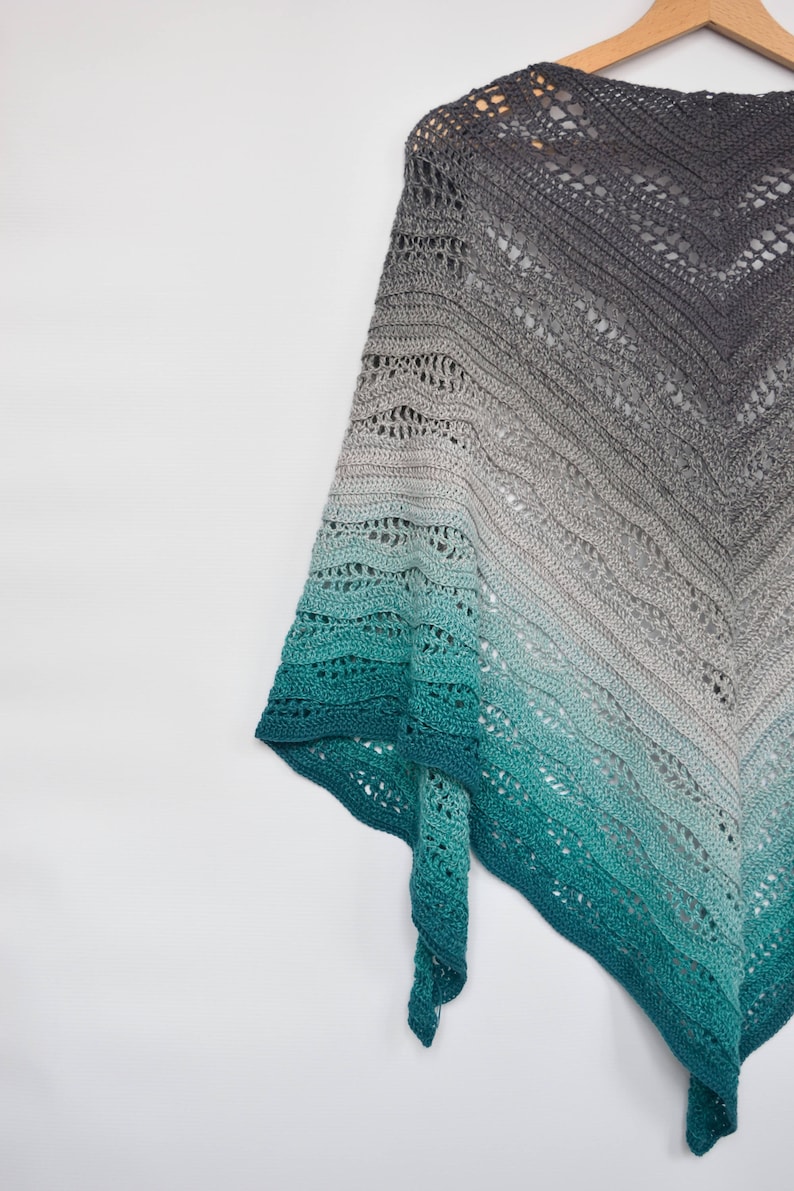 May include: A handmade, open-weave shawl in a gradient of colors, from dark gray to teal. The shawl is made of soft yarn and is draped over a wooden hanger. The intricate crochet pattern is visible throughout the design.