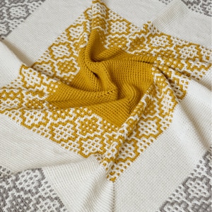 May include: A crocheted blanket with a geometric pattern in shades of white, gray, and yellow. The blanket is folded in half and has a textured surface.