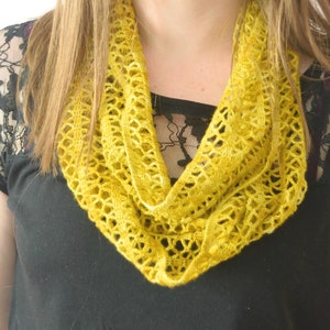 May include: A mustard yellow crocheted infinity scarf. The scarf is made of a delicate, open-weave pattern. The scarf is draped around the neck, and the bottom of the scarf forms a V-shape. The background is a black shirt.