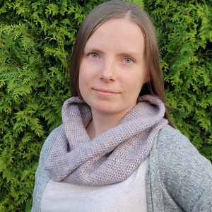 May include: A woman wearing a light purple crocheted infinity scarf. The scarf is made with a simple stitch pattern and has a relaxed, drapey look. The woman is wearing a white shirt and a gray cardigan.