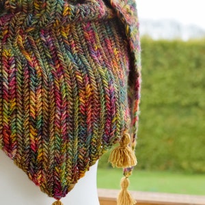 Może przedstawiać: Trójkątny szal wykonany na szydełku z kolorowej przędzy w odcieniach zieleni, brązu, czerwieni i żółci. Szal ma trzy frędzle na dolnych rogach.