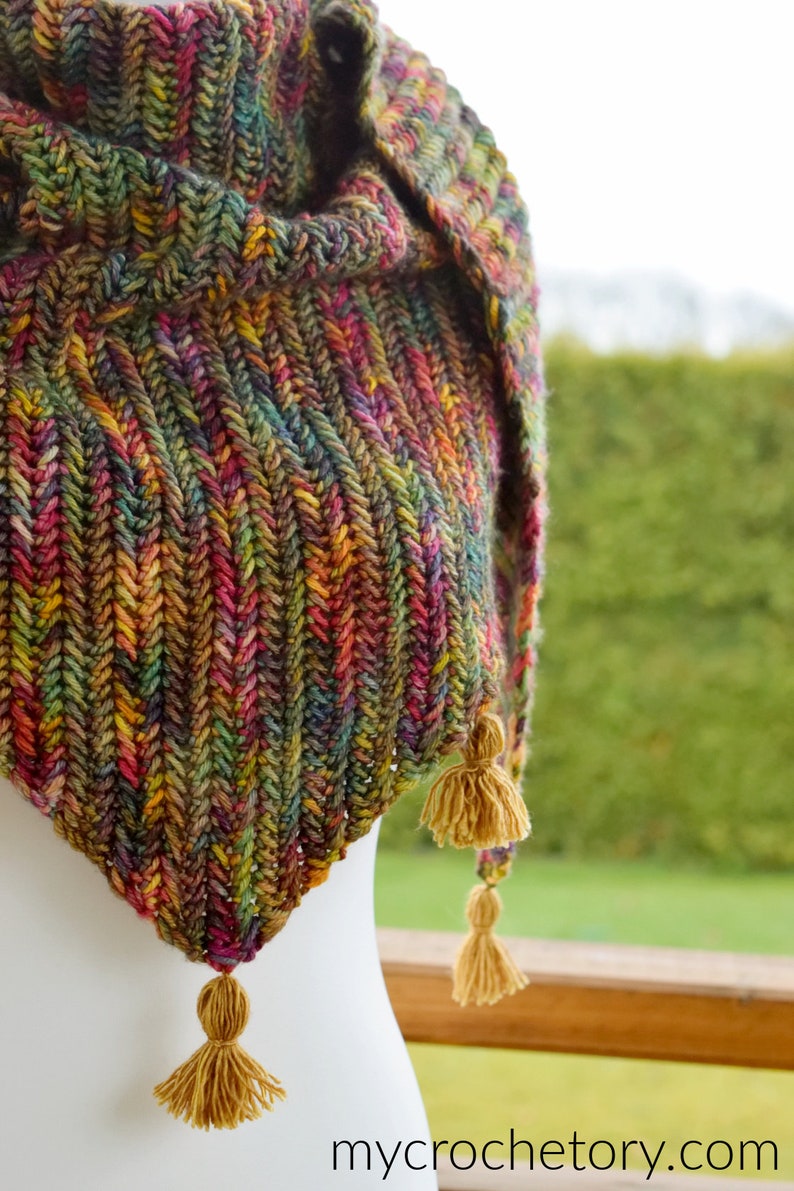 Puede incluir: Un chal de ganchillo triangular con un hilo de colores en tonos verdes, marrones, rojos y amarillos. El chal tiene tres borlas en las esquinas inferiores.