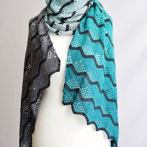 May include: A crocheted shawl with a chevron pattern in shades of teal, gray, and black. The shawl is draped over a white mannequin.
