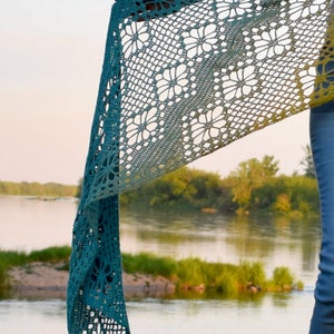 May include: A teal and yellow crochet shawl with a floral pattern. The shawl is draped over a person's arm and the background is a blurry image of a river and trees.