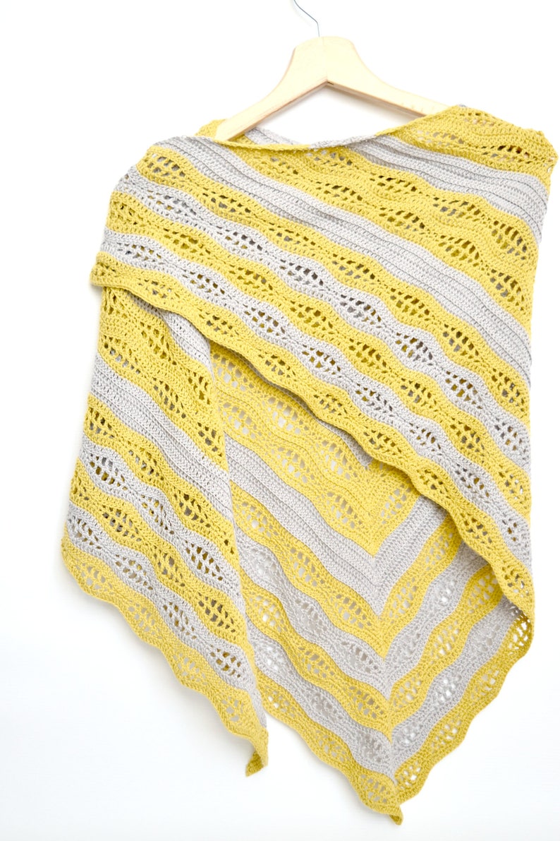 May include: A yellow and gray crocheted shawl with a striped pattern. The shawl is hanging on a wooden hanger against a white background. The design features alternating bands of yellow and gray, with a scalloped edge.