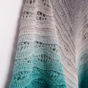 May include: A handmade crochet shawl with a gradient color scheme, transitioning from gray to teal. The shawl features an open, lacy design with intricate stitch patterns. The soft, textured fabric drapes elegantly, showcasing the craftsmanship.