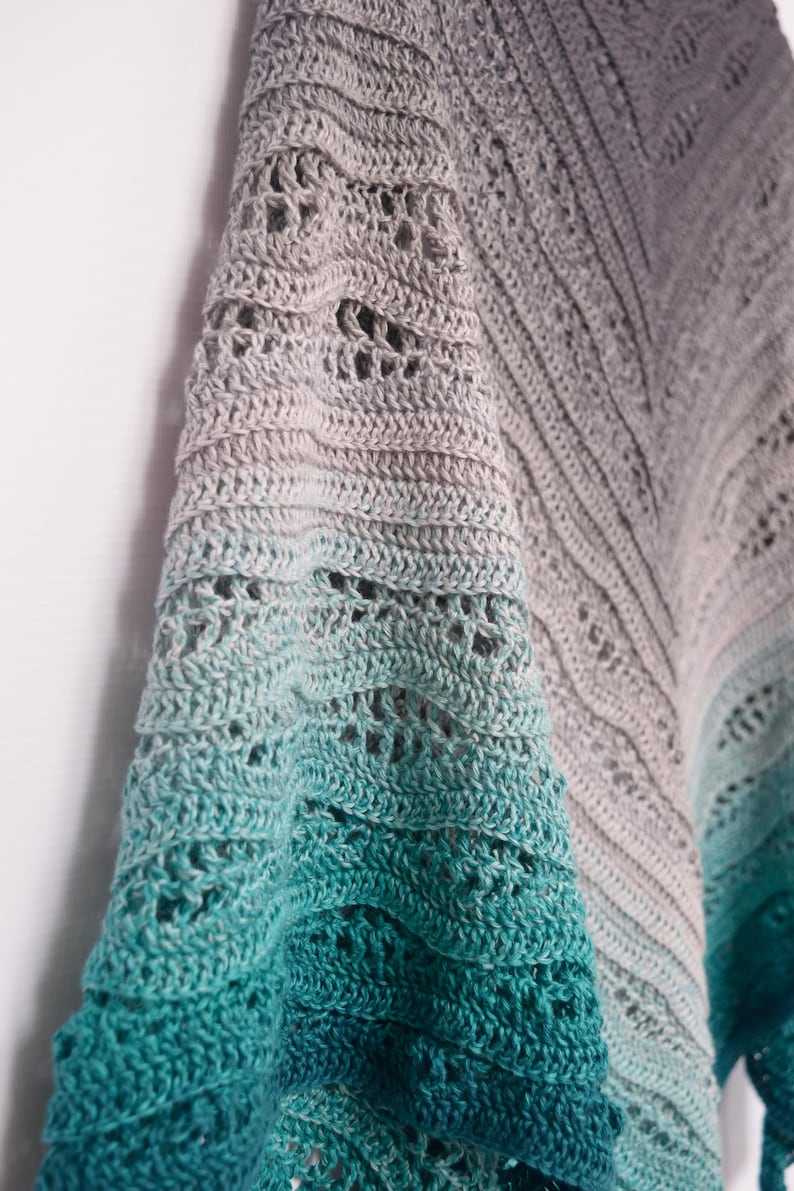 May include: A handmade crochet shawl with a gradient color scheme, transitioning from gray to teal. The shawl features an open, lacy design with intricate stitch patterns. The soft, textured fabric drapes elegantly, showcasing the craftsmanship.