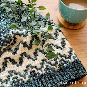 Peut inclure: Une couverture au crochet avec un motif géométrique vert et blanc. La couverture est pliée et drapée sur une surface en bois avec une plante verte et une tasse bleue sur un dessous-de-verre en bois.