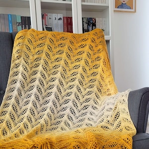 Może przedstawiać: Ręcznie robiony koc w kolorze ż&oacute;łtym i białym w ombre z wzorem chevron. Koc jest położony na szarym fotelu w salonie.