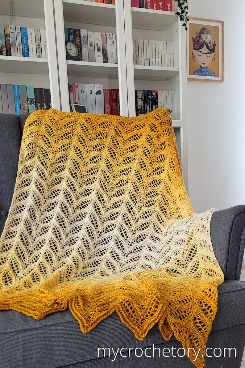 Może przedstawiać: Ręcznie robiony koc w kolorze ż&oacute;łtym i białym w ombre z wzorem chevron. Koc jest położony na szarym fotelu w salonie.