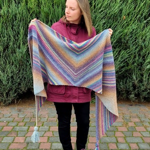 Peut inclure: Un châle crocheté à la main, multicolore, avec un design encolure en V. Le châle présente un dégradé de couleurs, notamment du bleu, du violet, du jaune et du marron. Il comporte des pompons aux extrémités des longs côtés. Le site web mycrochetory.com est visible en bas.