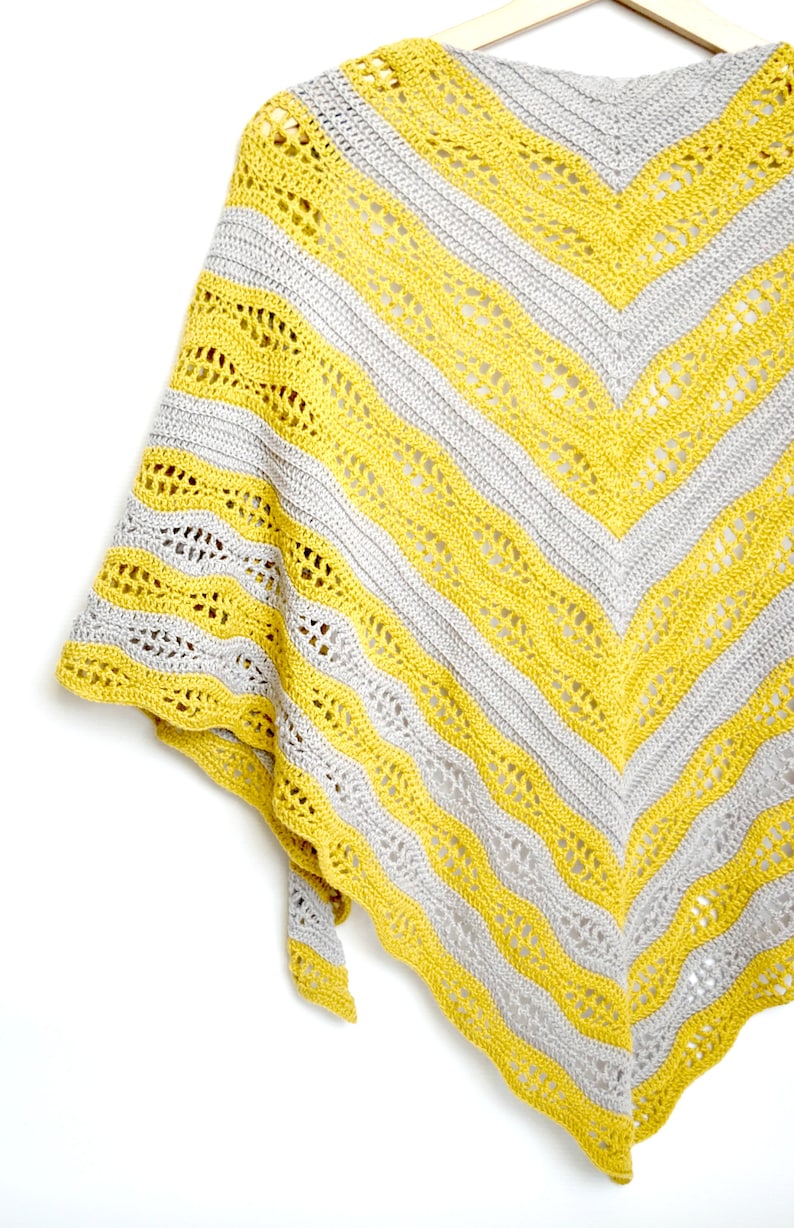 May include: A handmade crochet shawl in a chevron pattern, featuring alternating stripes of yellow and gray. The shawl is displayed on a wooden hanger against a white background, showcasing its intricate openwork design.