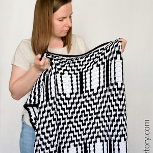 May include: A person holding a black and white crocheted blanket with a geometric pattern. The blanket has a zig-zag pattern and a series of rectangular shapes.