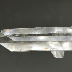 Può includere: Un cristallo di quarzo trasparente con doppia terminazione, che mostra striature interne e un leggero bagliore iridescente.