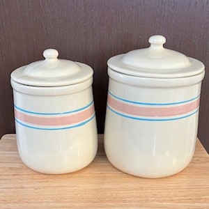 May include: Two off-white ceramic canisters with lids. Each canister has a pink and blue stripe around the middle. The canisters are on a wooden surface against a brown background.