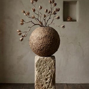 May include: A decorative, spherical vase with a textured, brown surface, holding dried branches with seed pods. The vase sits atop a weathered, rectangular stone pedestal, creating a rustic, natural aesthetic. The overall design evokes a sense of organic elegance.