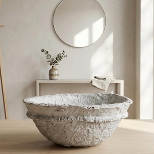 May include: A large, textured, off-white decorative bowl with a wide rim sits on a light wood surface. The bowl has a rough, organic appearance. A round mirror and a small vase with greenery are in the background, creating a minimalist aesthetic.