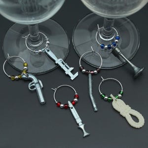 Peut inclure: Un assortiment de marque-verres à vin aux designs originaux. Les breloques comprennent un pistolet, une clé, un couteau, une corde et d'autres formes. Chaque breloque a un cerceau argenté avec des perles colorées. Les breloques sont présentées sur une surface sombre.