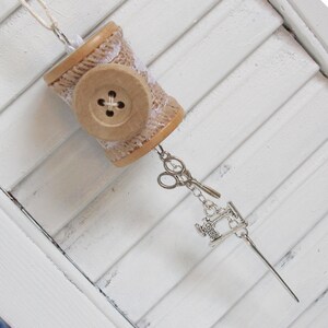 May include: A wooden spool wrapped in burlap and lace with a button and a charm of a sewing machine, scissors, and a needle hanging from it.
