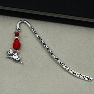 May include: Silver-toned metal bookmark with a decorative cat charm and red bead accents. The bookmark features a curved design with an etched pattern along the edge. A small, detailed cat charm dangles from the top, adding a whimsical touch.