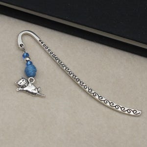 May include: Silver-toned metal bookmark with a decorative cat charm and blue beads. The bookmark has a curved design with an etched pattern along the edge. The cat charm is in mid-leap.