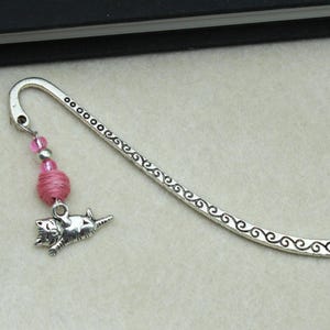 May include: Silver-toned metal bookmark with a curled top and a decorative swirl pattern. A pink beaded charm and a small silver cat charm dangle from the top. The cat is lying down.
