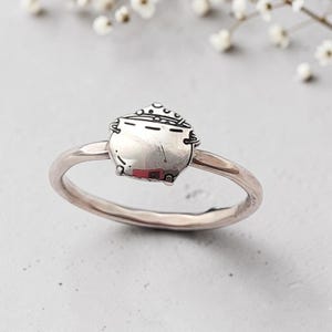 May include: A silver-toned ring featuring a small, detailed charm shaped like a cauldron. The cauldron charm has a smooth, rounded body with etched lines along the top rim and sides, and the ring band is a simple, polished, thin metal loop.