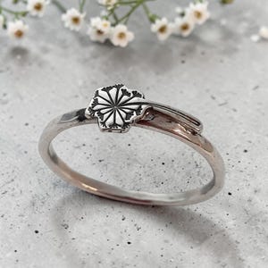 Peut inclure: Une bague argentée ornée d'un motif plat et circulaire représentant une tête de pissenlit avec des lignes rayonnantes. L'anneau est lisse et poli, le motif floral s'étendant légèrement sur le bord supérieur pour un aspect détaillé et délicat.