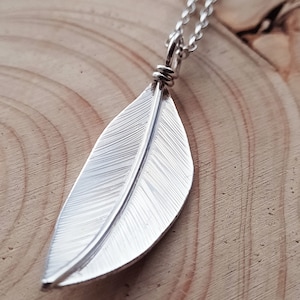 Puede incluir: Un collar con un colgante de pluma de plata. La pluma está detallada con líneas finas y cuelga de una cadena de plata.