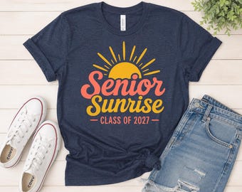 Könnte beinhalten: Marineblaues T-Shirt mit einem gelben Sonnensymbol und den Worten "Senior Sunrise" in Koralle und Gelb, darunter "Class of 2027". Das Shirt wird mit Jeansshorts und weißen Turnschuhen kombiniert.