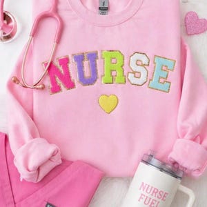 May include: A pink sweatshirt with the word "NURSE" in colorful, glittery letters. A yellow heart is below the word. A pink stethoscope, a tumbler with "NURSE FUEL" text, and pink scrubs are also in the image.