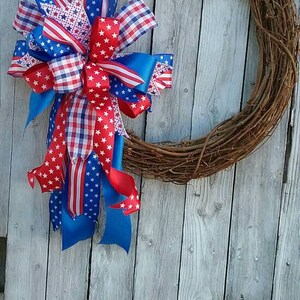 May include: A grapevine wreath decorated with a large bow made of red, white, and blue ribbon. The ribbon features a stars and stripes pattern, gingham, and solid colors.