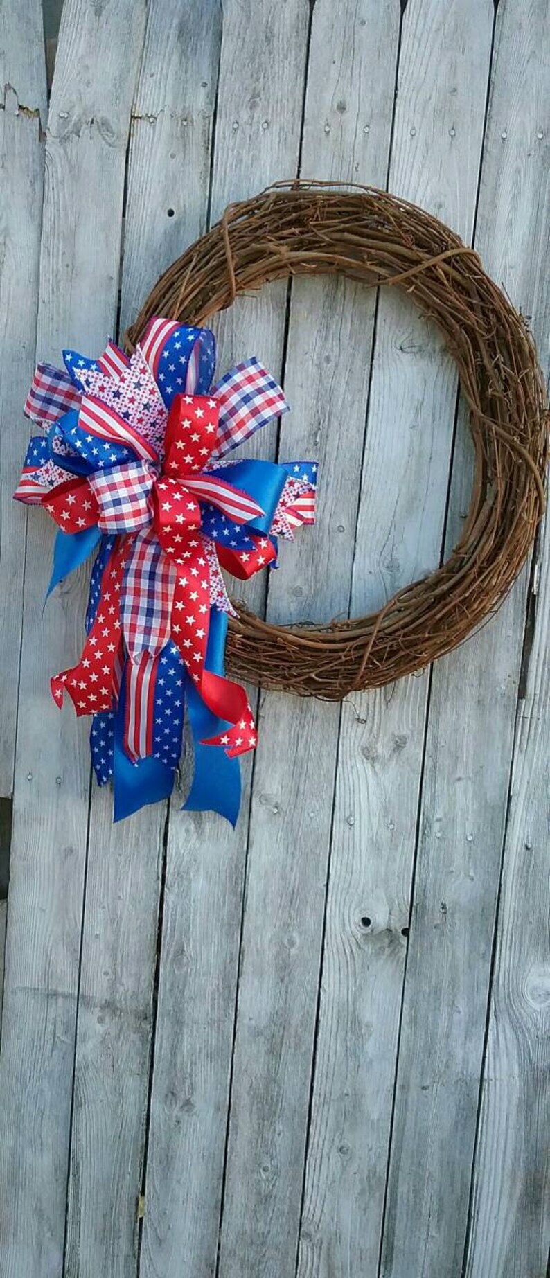 May include: A grapevine wreath decorated with a large bow made of red, white, and blue ribbon. The ribbon features a stars and stripes pattern, gingham, and solid colors.