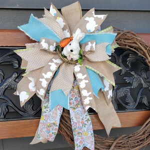 May include: Easter-themed bow with a white bunny in a nest, surrounded by ribbons in various patterns and colors. The bow features burlap, polka dots, and bunny prints, with accents of blue and floral patterns. A decorative wreath is visible.