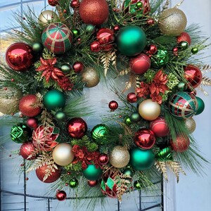 May include: A Christmas wreath decorated with red, green, and gold ornaments. The wreath is made of pine branches and has a variety of different ornaments, including balls, stars, and bows.