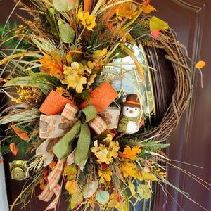 May include: A fall-themed wreath with a snowman figurine, featuring a variety of autumnal colors and textures, including orange, brown, green, and yellow. The wreath is decorated with leaves, berries, and grasses.
