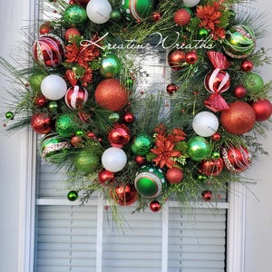 May include: A Christmas wreath featuring green pine needles, red and green ornaments, white spheres, and red poinsettia flowers. The wreath includes candy-shaped decorations and glittery accents, with the text Kreaten Wreaths in the center.