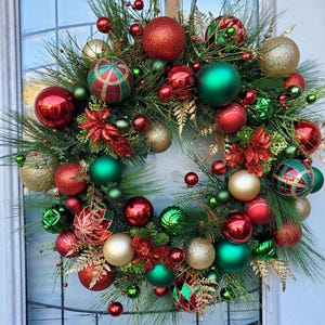 May include: A Christmas wreath decorated with red, green, and gold ornaments. The wreath is made of pine branches and has a variety of different ornaments, including round balls, striped balls, and star-shaped ornaments.