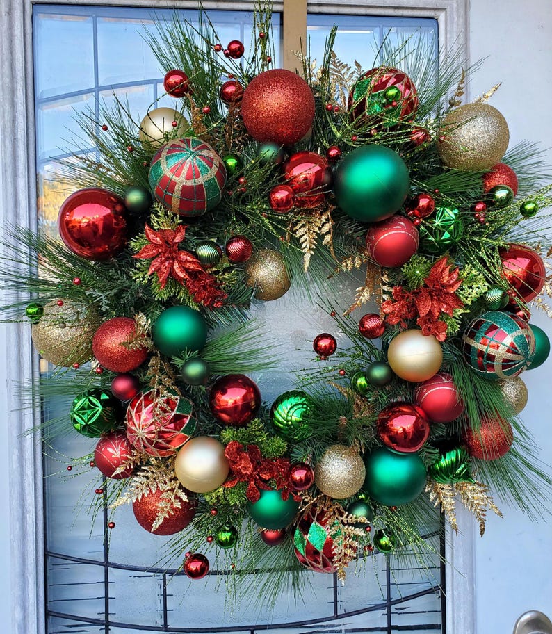 May include: A Christmas wreath decorated with red, green, and gold ornaments. The wreath is made of pine branches and has a variety of different ornaments, including round balls, striped balls, and star-shaped ornaments.