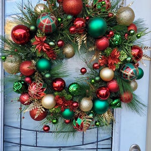 May include: A Christmas wreath decorated with red, green, and gold ornaments. The wreath is made of pine branches and has a variety of different ornaments, including balls, stars, and bows.