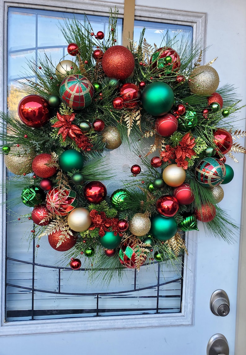 May include: A Christmas wreath decorated with red, green, and gold ornaments. The wreath is made of pine branches and has a variety of different ornaments, including balls, stars, and bows.