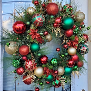 May include: A Christmas wreath decorated with red, green, and gold ornaments. The wreath is made of pine branches and has a variety of different ornaments, including round ornaments, star ornaments, and ribbon ornaments.